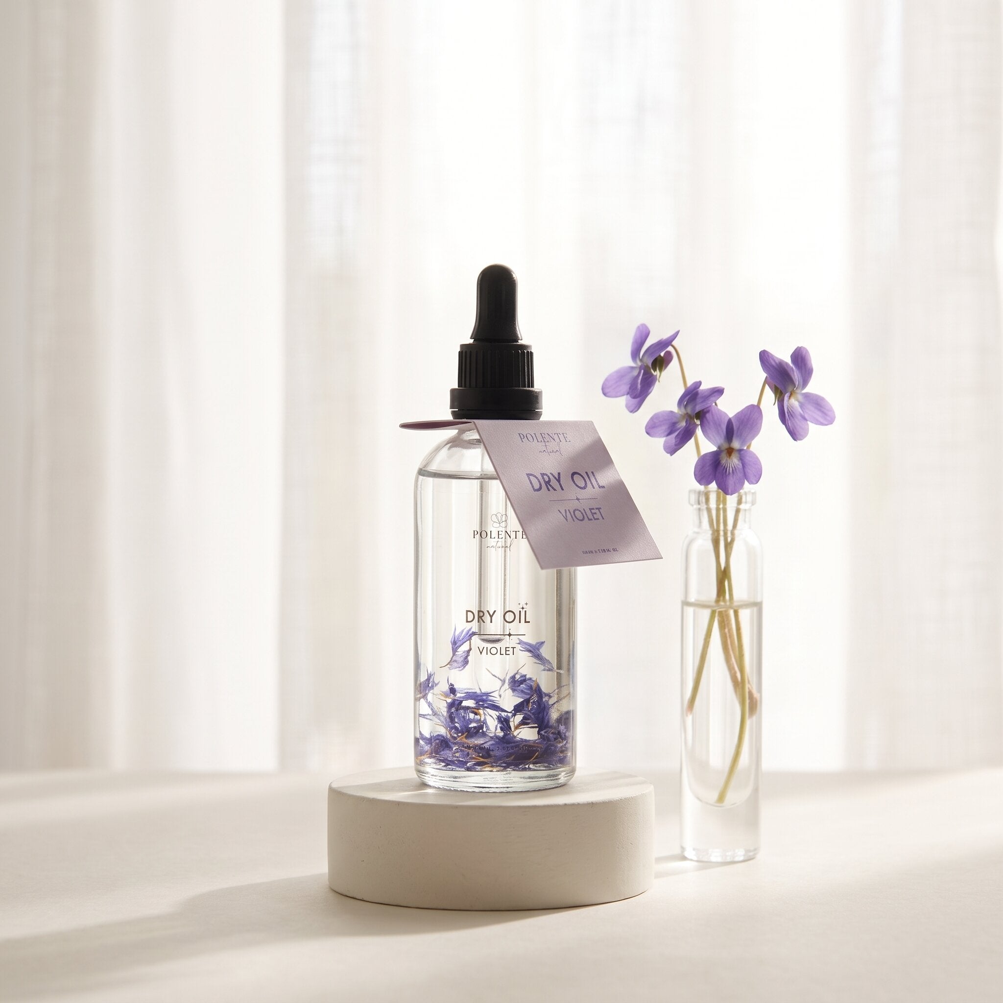 Violet Dry Oil 100 ml çok amaçlı kuru yağ — beyaz sıva kaide ve tül perde ışığında menekşeli konsept görseli