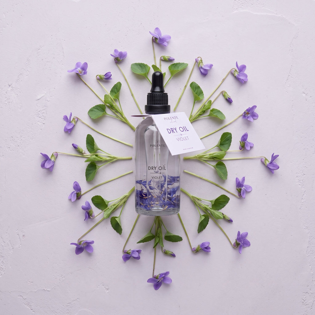 Violet Dry Oil 100 ml çok amaçlı kuru yağ — açık lila sıva üzerinde menekşe dallarıyla simetrik konsept görseli
