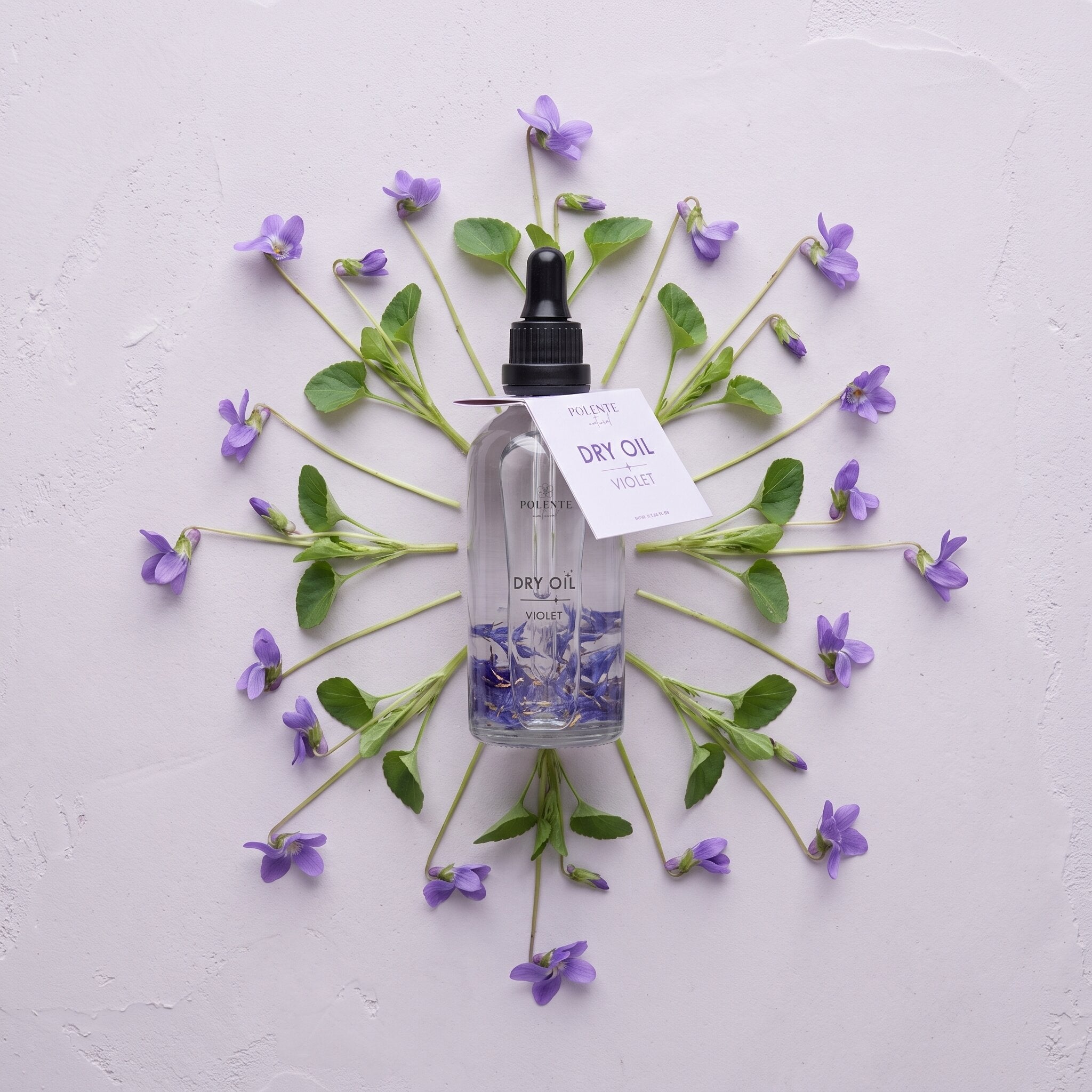 Violet Dry Oil 100 ml çok amaçlı kuru yağ — açık lila sıva üzerinde menekşe dallarıyla simetrik konsept görseli