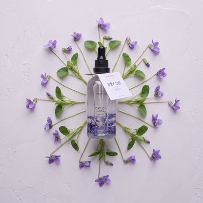Violet Dry Oil 100 ml çok amaçlı kuru yağ — açık lila sıva üzerinde menekşe dallarıyla simetrik konsept görseli
