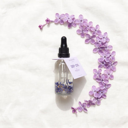 Violet Dry Oil 50 ml çok amaçlı kuru yağ — fildişi keten üzerinde menekşe yapraklarıyla botanik konsept görseli