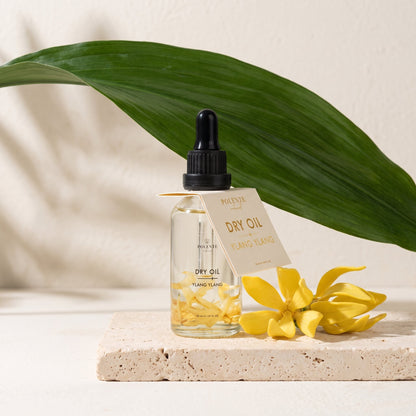 Ylang Ylang Dry Oil 50 ml çok amaçlı kuru yağ — travertem üzerinde tropikal yaprak ve ylang ylang çiçekleriyle konsept görseli